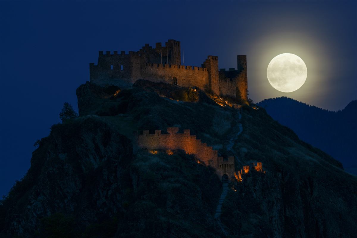 A full moon rising behind a floodlit stone castle on a dark hill